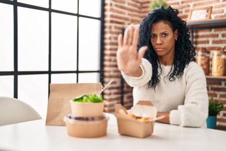 Photo for Middle age hispanic woman eating take away food with open hand doing stop sign with serious and confident expression, defense gesture - Royalty Free Image