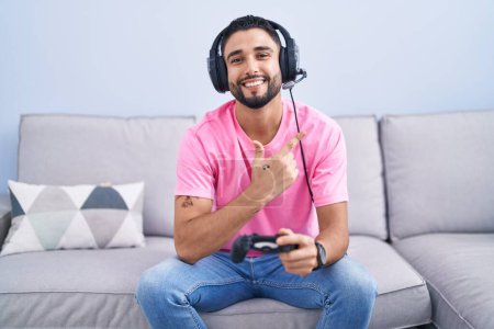 Photo for Hispanic young man playing video game holding controller sitting on the sofa cheerful with a smile on face pointing with hand and finger up to the side with happy and natural expression - Royalty Free Image