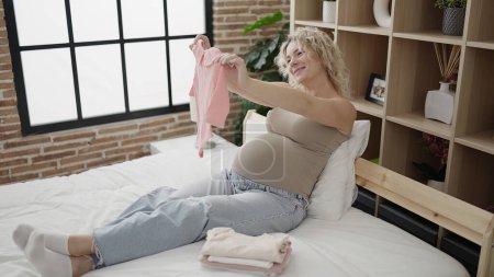 Téléchargez les photos : Jeune femme enceinte tenant des vêtements de bébé assis sur le lit dans la chambre - en image libre de droit