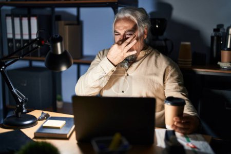Téléchargez les photos : Homme d'âge moyen aux cheveux gris travaillant au bureau la nuit sentant quelque chose de puant et dégoûtant, odeur intolérable, retenant son souffle avec les doigts sur le nez. mauvaise odeur - en image libre de droit