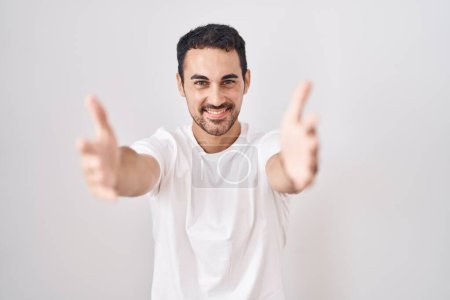 Photo for Handsome hispanic man standing over white background looking at the camera smiling with open arms for hug. cheerful expression embracing happiness. - Royalty Free Image
