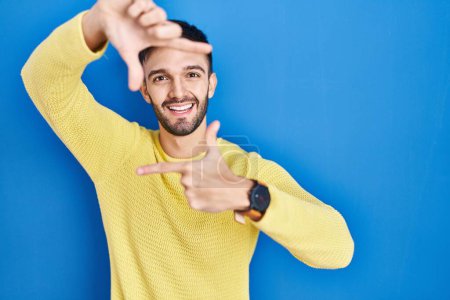 Téléchargez les photos : Hispanic man standing over blue background smiling making frame with hands and fingers with happy face. creativity and photography concept. - en image libre de droit