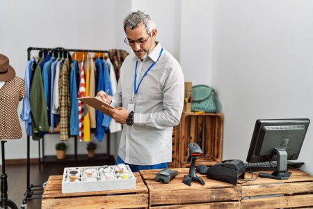 Foto de Hombre de pelo gris de mediana edad asistente de la tienda de escribir en el documento mirando reloj en la tienda de ropa - Imagen libre de derechos