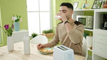 Téléchargez les photos : Jeune homme arabe boire un verre de lait assis sur la table à la maison - en image libre de droit