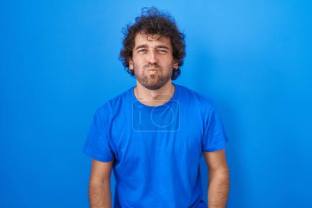 Foto de Hispanic young man standing over blue background puffing cheeks with funny face. mouth inflated with air, crazy expression. - Imagen libre de derechos