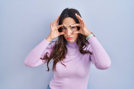 Téléchargez les photos : Jeune femme brune debout sur fond bleu essayant d'ouvrir les yeux avec les doigts, endormie et fatiguée pour la fatigue matinale - en image libre de droit