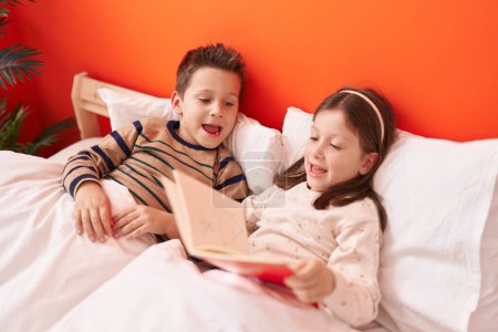 Photo for Adorable boy and girl reading book sitting on bed at bedroom - Royalty Free Image