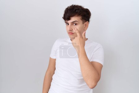 Téléchargez les photos : Jeune homme non binaire portant un t-shirt blanc décontracté pointant vers l'œil vous regardant geste, expression suspecte - en image libre de droit