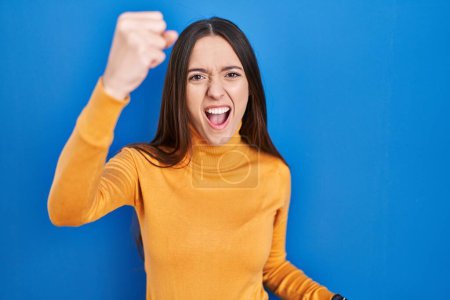 Photo for Young brunette woman standing over blue background angry and mad raising fist frustrated and furious while shouting with anger. rage and aggressive concept. - Royalty Free Image