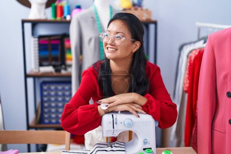 Foto de Joven mujer china sastre sonriendo confiado apoyándose en la máquina de coser en atelier - Imagen libre de derechos