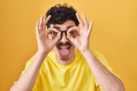 Foto de Hombre hispano con gafas de pie sobre fondo amarillo haciendo buen gesto como prismáticos sacando la lengua, ojos mirando a través de los dedos. expresión loca. - Imagen libre de derechos