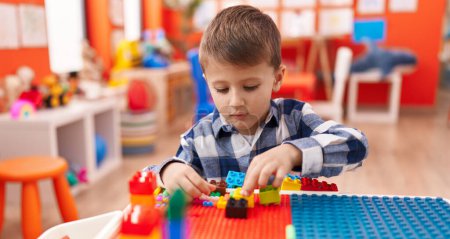 Foto de Adorable chico caucásico jugando con bloques de construcción sentado en la mesa en el jardín de infantes - Imagen libre de derechos