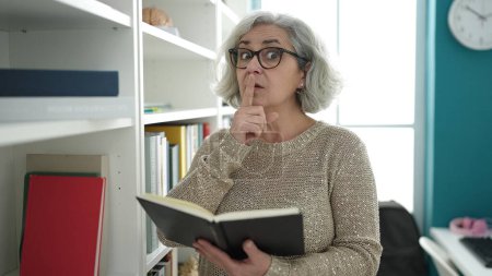 Téléchargez les photos : Femme d'âge moyen avec professeur de cheveux gris lisant un livre faisant un geste de silence dans une salle de classe universitaire - en image libre de droit