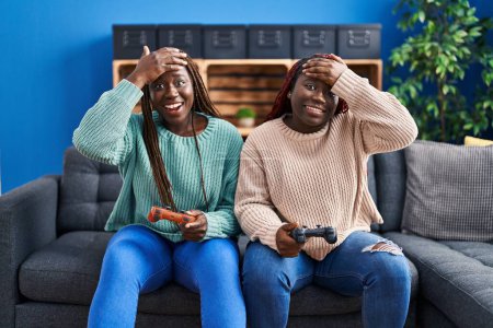 Foto de Dos mujeres africanas jugando videojuego sosteniendo controlador estresado y frustrado con la mano en la cabeza, cara sorprendida y enojada - Imagen libre de derechos