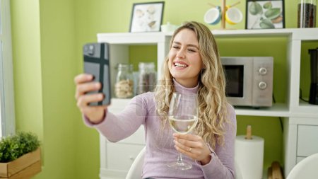 Foto de Joven rubia sosteniendo copa de vino teniendo videollamada en el comedor - Imagen libre de derechos