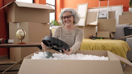 Téléchargez les photos : Middle age woman with grey hair smiling confident unpacking cardboard box at new home - en image libre de droit