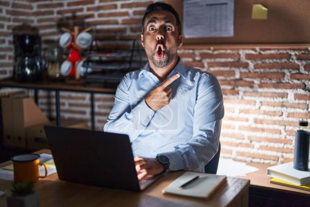 Photo for Hispanic man with beard working at the office at night surprised pointing with finger to the side, open mouth amazed expression. - Royalty Free Image