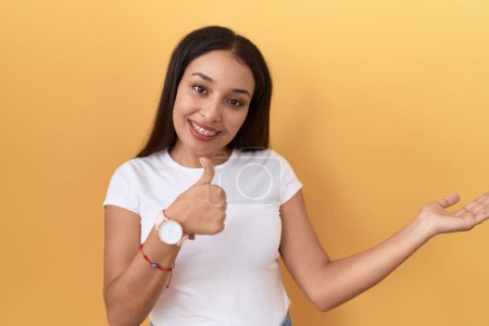 Photo for Young arab woman wearing casual white t shirt over yellow background showing palm hand and doing ok gesture with thumbs up, smiling happy and cheerful - Royalty Free Image