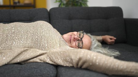 Téléchargez les photos : Femme d'âge moyen avec les cheveux gris couché sur le canapé détendu à la maison - en image libre de droit