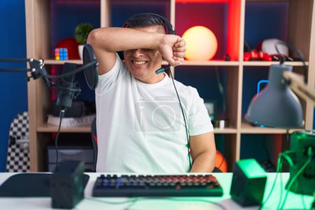 Téléchargez les photos : Jeune homme hispanique jouant à des jeux vidéo couvrant les yeux avec le bras souriant gai et drôle. concept aveugle. - en image libre de droit