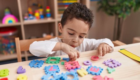 Téléchargez les photos : Adorable garçon hispanique jouant avec le jeu de puzzle de vocabulaire assis sur la table à la maternelle - en image libre de droit
