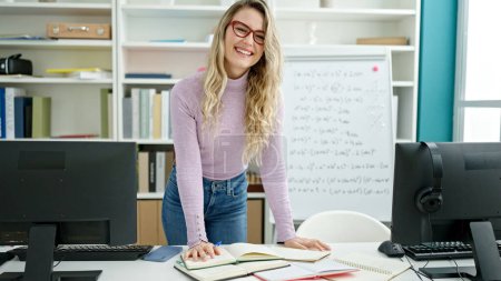Téléchargez les photos : Jeune enseignante blonde souriante écriture confiante sur carnet de notes en classe - en image libre de droit
