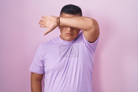 Foto de Joven hispano de pie sobre fondo rosa cubriendo los ojos con el brazo, luciendo serio y triste. concepto de ocultamiento y rechazo sin visión - Imagen libre de derechos