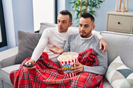 Photo for Homosexual couple eating popcorn watching tv clueless and confused expression. doubt concept. - Royalty Free Image
