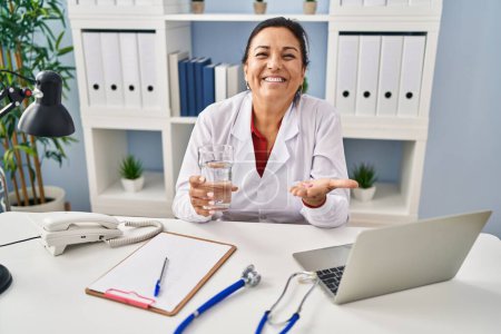 Foto de Médico maduro hispano mujer sosteniendo píldora y vaso de agua sonriendo y riendo en voz alta porque broma loca divertida. - Imagen libre de derechos