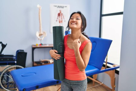 Photo for Young asian woman holding yoga mat at clinic screaming proud, celebrating victory and success very excited with raised arm - Royalty Free Image