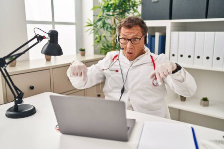 Foto de Hombre médico senior que trabaja en una cita en línea apuntando hacia abajo con los dedos mostrando publicidad, cara sorprendida y boca abierta - Imagen libre de derechos