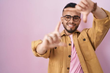 Téléchargez les photos : Jeune homme hispanique debout sur fond rose souriant faisant cadre avec les mains et les doigts avec le visage heureux. concept de créativité et photographie. - en image libre de droit