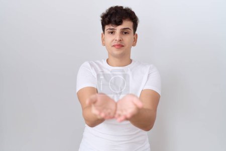 Téléchargez les photos : Jeune homme non binaire portant un t-shirt blanc décontracté souriant avec les mains paumes ensemble recevant ou donnant geste. maintien et protection - en image libre de droit