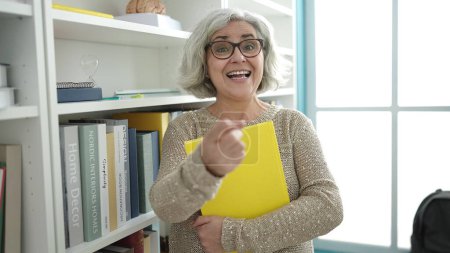 Photo for Middle age woman with grey hair teacher holding book pointing with finger at university classroom - Royalty Free Image