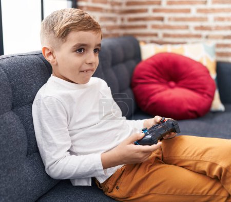 Foto de Adorable chico hispano jugando videojuego sentado en el sofá en casa - Imagen libre de derechos