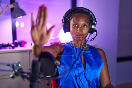 Foto de Mujer africana con rastas usando auriculares con la mano abierta haciendo stop sign con expresión seria y segura, gesto de defensa - Imagen libre de derechos