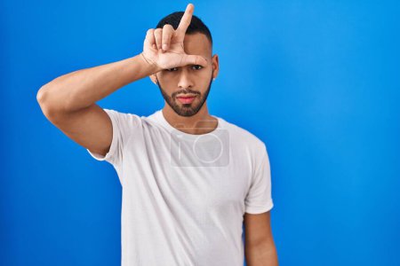 Téléchargez les photos : Young hispanic man standing over blue background making fun of people with fingers on forehead doing loser gesture mocking and insulting. - en image libre de droit