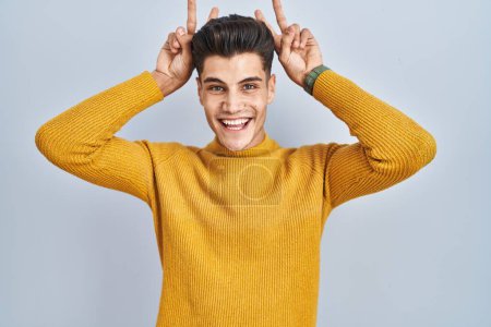 Foto de Joven hombre hispano de pie sobre fondo azul posando divertido y loco con los dedos en la cabeza como orejas de conejo, sonriendo alegre - Imagen libre de derechos