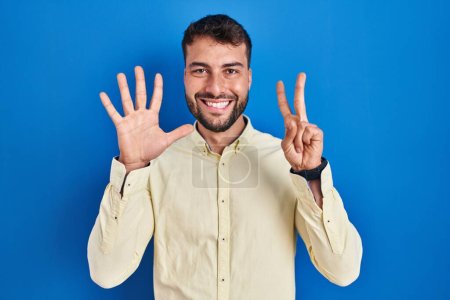 Téléchargez les photos : Bel homme hispanique debout sur fond bleu montrant et pointant vers le haut avec les doigts numéro sept tout en souriant confiant et heureux. - en image libre de droit