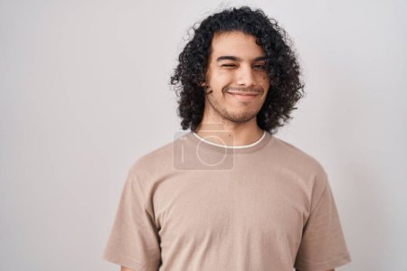 Téléchargez les photos : Homme hispanique avec les cheveux bouclés debout sur fond blanc clin d'oeil en regardant la caméra avec une expression sexy, gai et heureux visage. - en image libre de droit