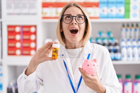 Foto de Joven mujer caucásica trabajando en farmacia sosteniendo píldoras una alcancía enojada y loca gritando frustrada y furiosa, gritando con ira mirando hacia arriba. - Imagen libre de derechos