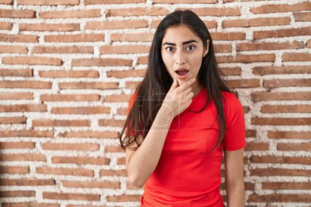 Photo for Young teenager girl standing over bricks wall looking fascinated with disbelief, surprise and amazed expression with hands on chin - Royalty Free Image