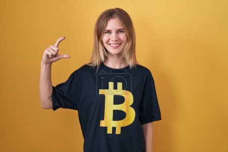 Téléchargez les photos : Femme caucasienne blonde portant Bitcoin t-shirt sourire et geste confiant avec la main faisant signe de petite taille avec les doigts à la recherche et la caméra. concept de mesure. - en image libre de droit