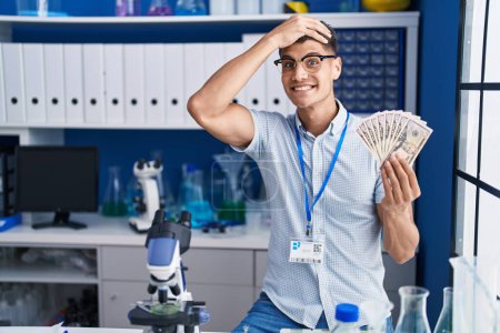 Foto de Joven hispano trabajando en laboratorio científico sosteniendo dólares estresados y frustrados con la mano en la cabeza, rostro sorprendido y enojado - Imagen libre de derechos