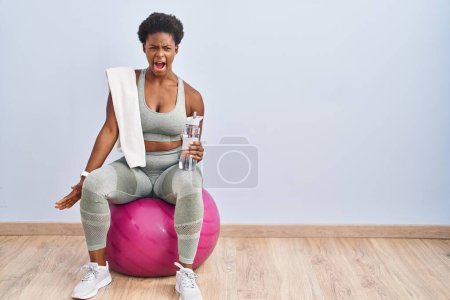 Téléchargez les photos : Femme afro-américaine portant des vêtements de sport assis sur pilates ballon en colère et fou hurlant frustré et furieux, criant de colère. rage et concept agressif. - en image libre de droit