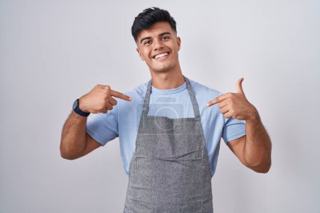 Photo for Hispanic young man wearing apron over white background looking confident with smile on face, pointing oneself with fingers proud and happy. - Royalty Free Image