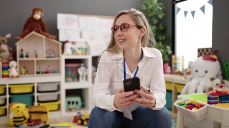 Photo for Young blonde woman preschool teacher smiling confident using smartphone at kindergarten - Royalty Free Image