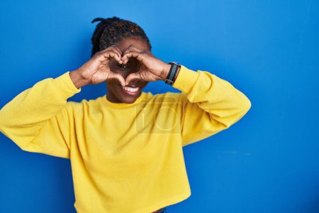 Photo for Beautiful black woman standing over blue background doing heart shape with hand and fingers smiling looking through sign - Royalty Free Image