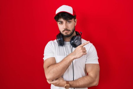 Téléchargez les photos : Homme hispanique avec barbe portant chapeau de joueur et écouteurs pointant avec le doigt de la main sur le côté montrant la publicité, visage sérieux et calme - en image libre de droit