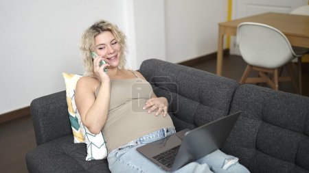 Photo for Young pregnant woman talking on smartphone using laptop touching belly at home - Royalty Free Image